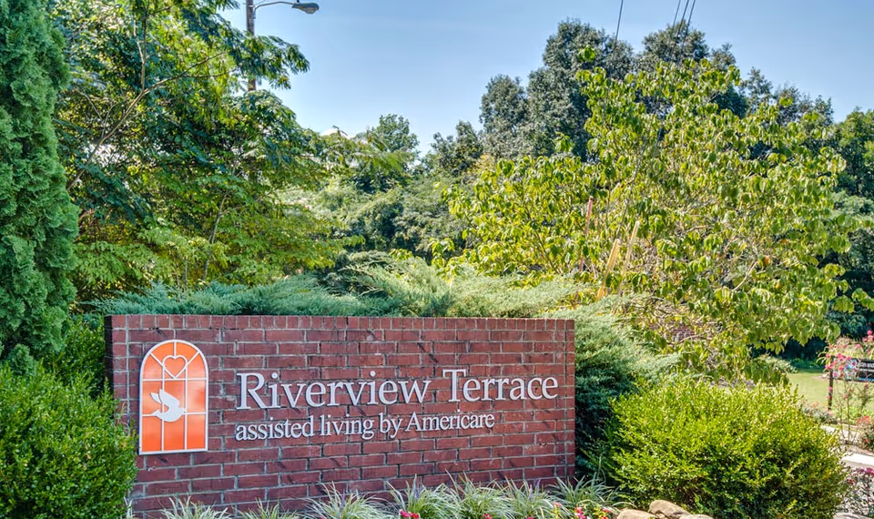 A brick sign for Riverview Terrace assisted living by Americare surrounded by green bushes and trees under a clear blue sky.