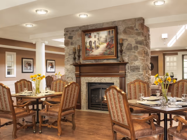 A cozy dining area with wooden tables and upholstered chairs arranged around a stone fireplace. The fireplace has a wooden mantel with decorative candlesticks and a framed painting above it. Vases with yellow flowers are placed on the tables. The room has warm lighting and a comfortable, inviting atmosphere.