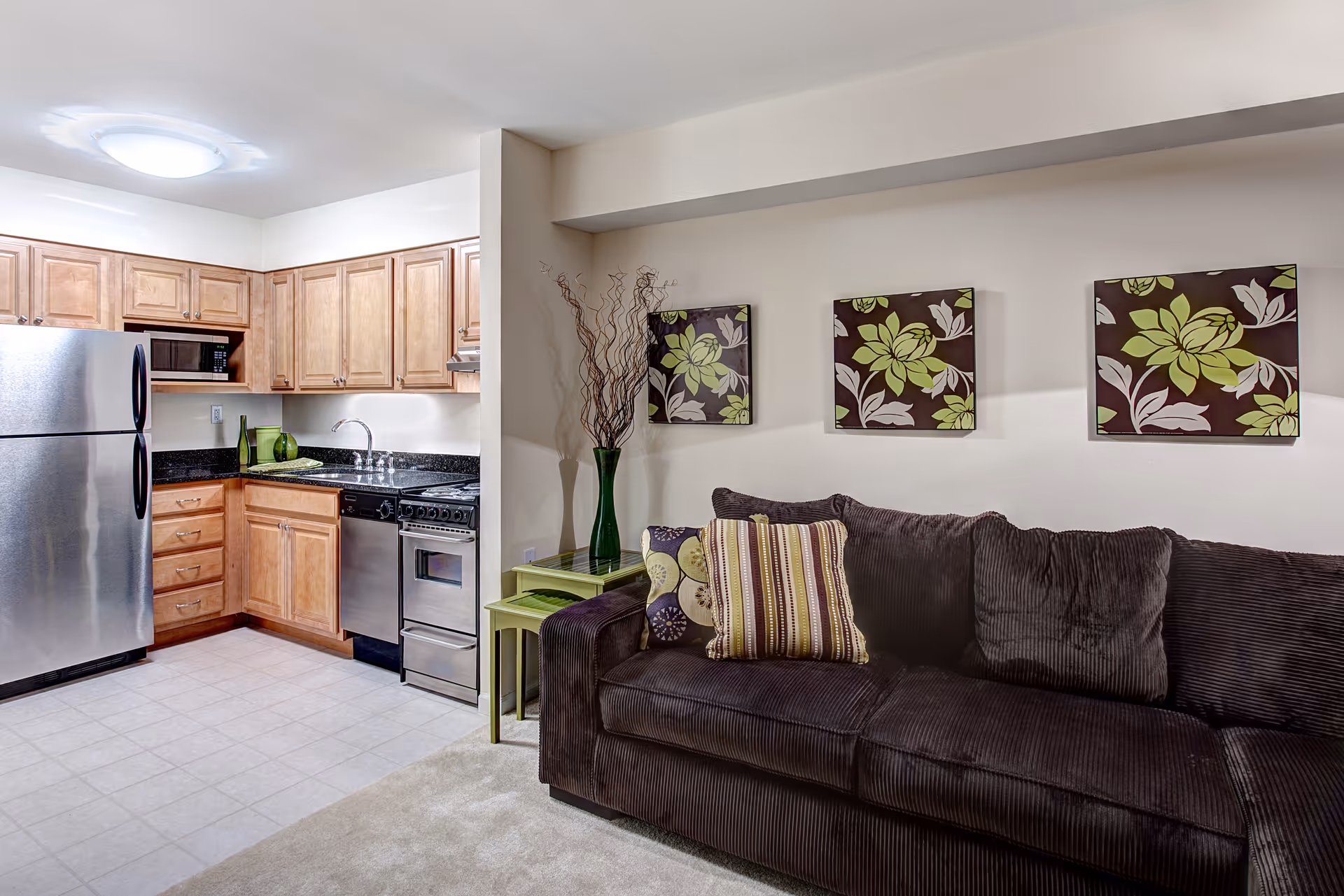 Interior view of a small living area and kitchen in a senior living facility. The kitchen features wooden cabinets, a stainless steel refrigerator, microwave, stove, and dishwasher. Adjacent to the kitchen is a dark brown couch with decorative pillows, a green side table with a tall green vase holding decorative branches, and three floral wall art pieces above the couch.