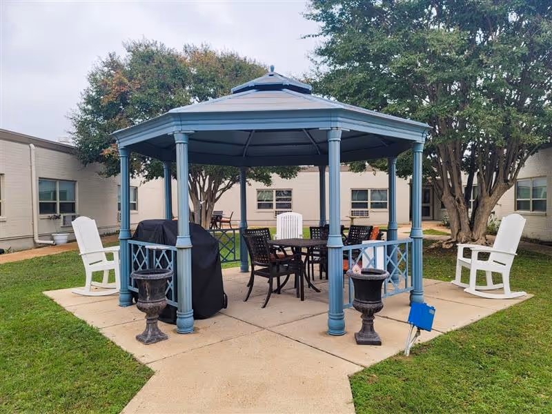 Outdoor gazebo with a blue roof and columns situated on a concrete patio surrounded by grass. Inside the gazebo are a round table and several chairs, with two white rocking chairs placed outside on either side. Trees and a beige building with windows are visible in the background.