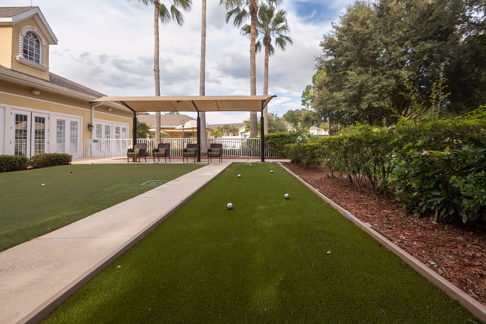 Outdoor bocce ball court with several bocce balls on the green turf, surrounded by bushes and trees. There is a covered seating area with chairs near a yellow building with white-framed windows and doors. Tall palm trees are visible in the background under a partly cloudy sky.