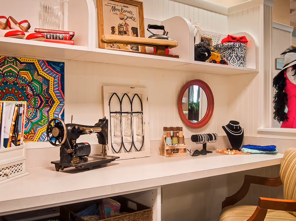 A craft or sewing room with a vintage sewing machine on a white countertop. The countertop also holds a small rack of colorful thread spools, jewelry displays with bracelets and necklaces, a round red-framed mirror, and various decorative items. Above the counter are white shelves with books, a framed picture, an old iron, and storage boxes. A colorful mandala artwork is partially visible on the left wall. A striped armchair is positioned at the counter.