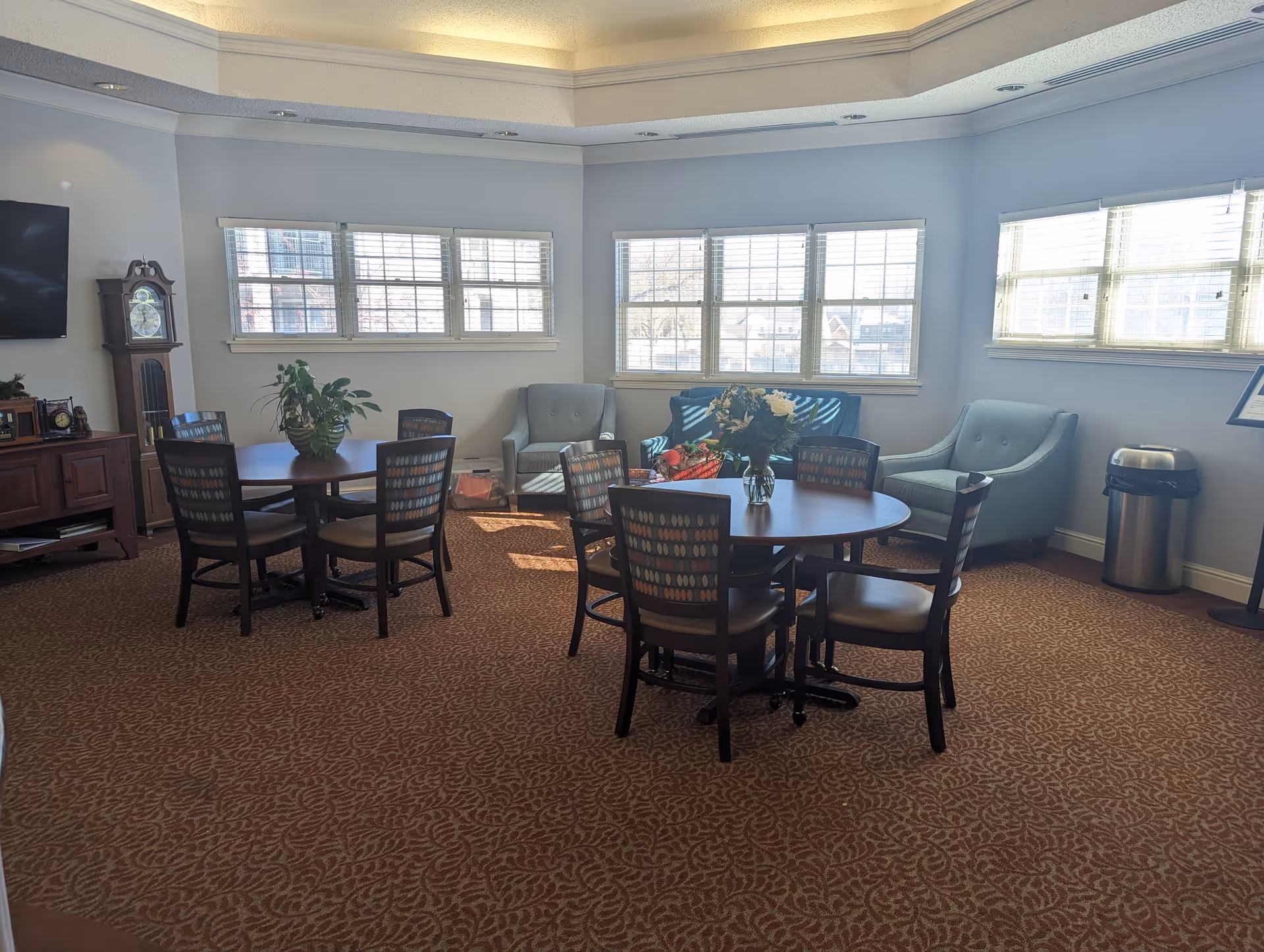 A well-lit common area with two round tables each surrounded by four chairs, a grandfather clock, a TV mounted on the wall, two armchairs, a loveseat, and large windows letting in natural light. There are plants and flowers on the tables, and a trash can in the corner.