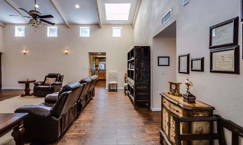 Interior view of a living area in an assisted living facility with leather recliner chairs arranged in a row, a ceiling fan with lights, small windows near the ceiling, wooden flooring, a decorative cabinet with framed certificates on the wall, and a bookshelf near an open doorway leading to a kitchen.