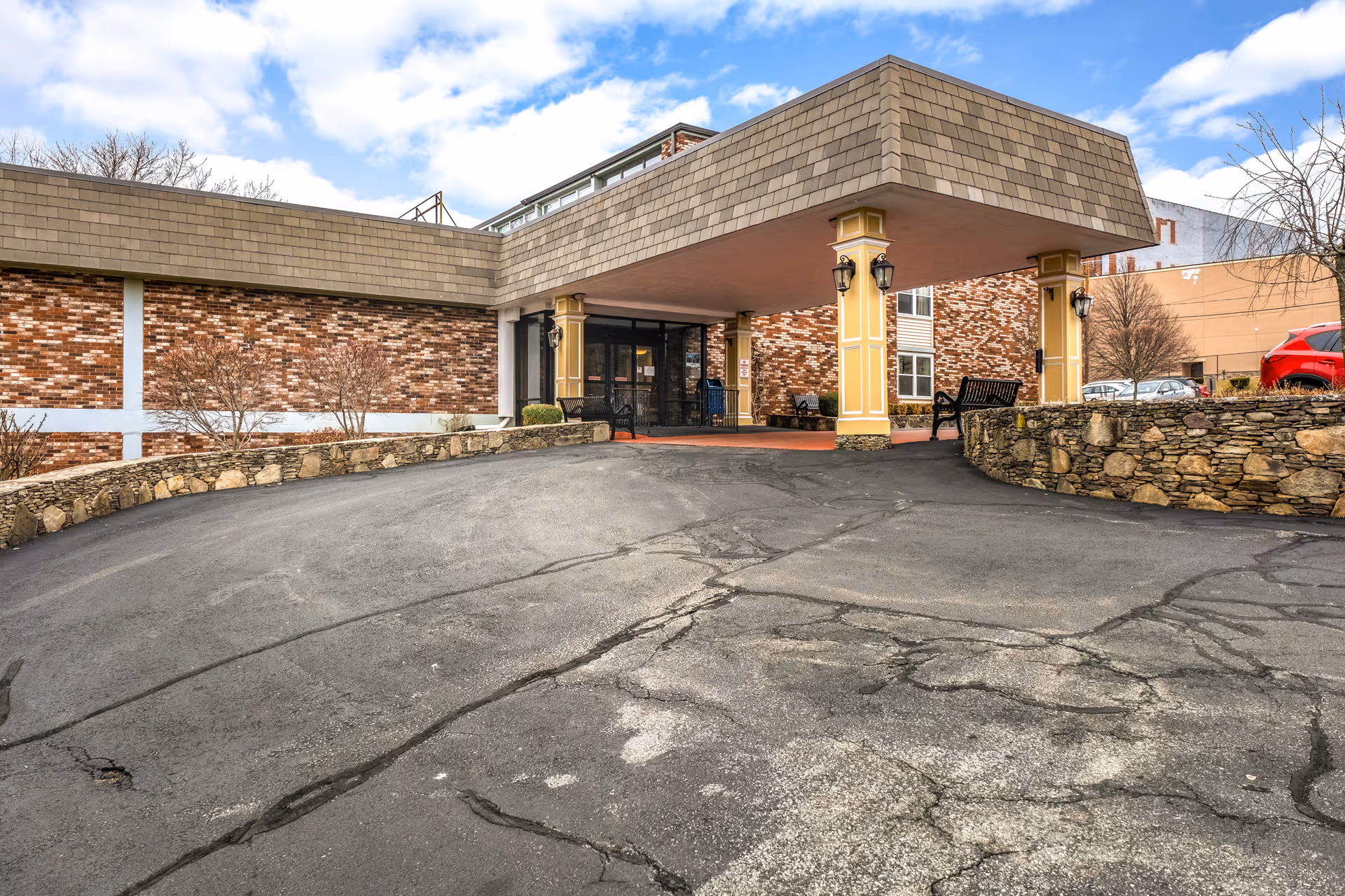 Covered entrance and porte-cochère of a brick senior living building with stone retaining walls, benches, and an asphalt driveway.