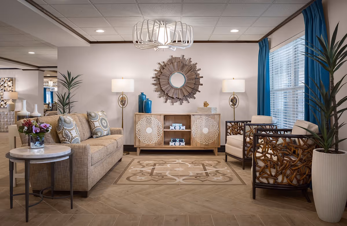 A well-decorated living room area with a beige sofa adorned with patterned cushions, two armchairs with wooden lattice designs, a round side table with a flower vase, a wooden cabinet with decorative items, two floor lamps, a large circular wooden wall mirror, blue curtains on a window, and two tall potted plants.