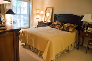 A cozy bedroom with a bed covered in a beige quilt and floral pillows. The room features a dark wooden headboard, a dresser with a lamp, a mirror, framed pictures on the wall, and a window with white blinds letting in natural light.