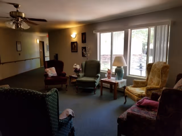 A cozy common area in a senior living facility with several upholstered armchairs arranged around a small wooden table holding a lamp and decorative items. Large windows let in natural light and offer a view of an outdoor space with a fountain. The room has carpeted floors, soft lighting, and framed pictures on the walls.