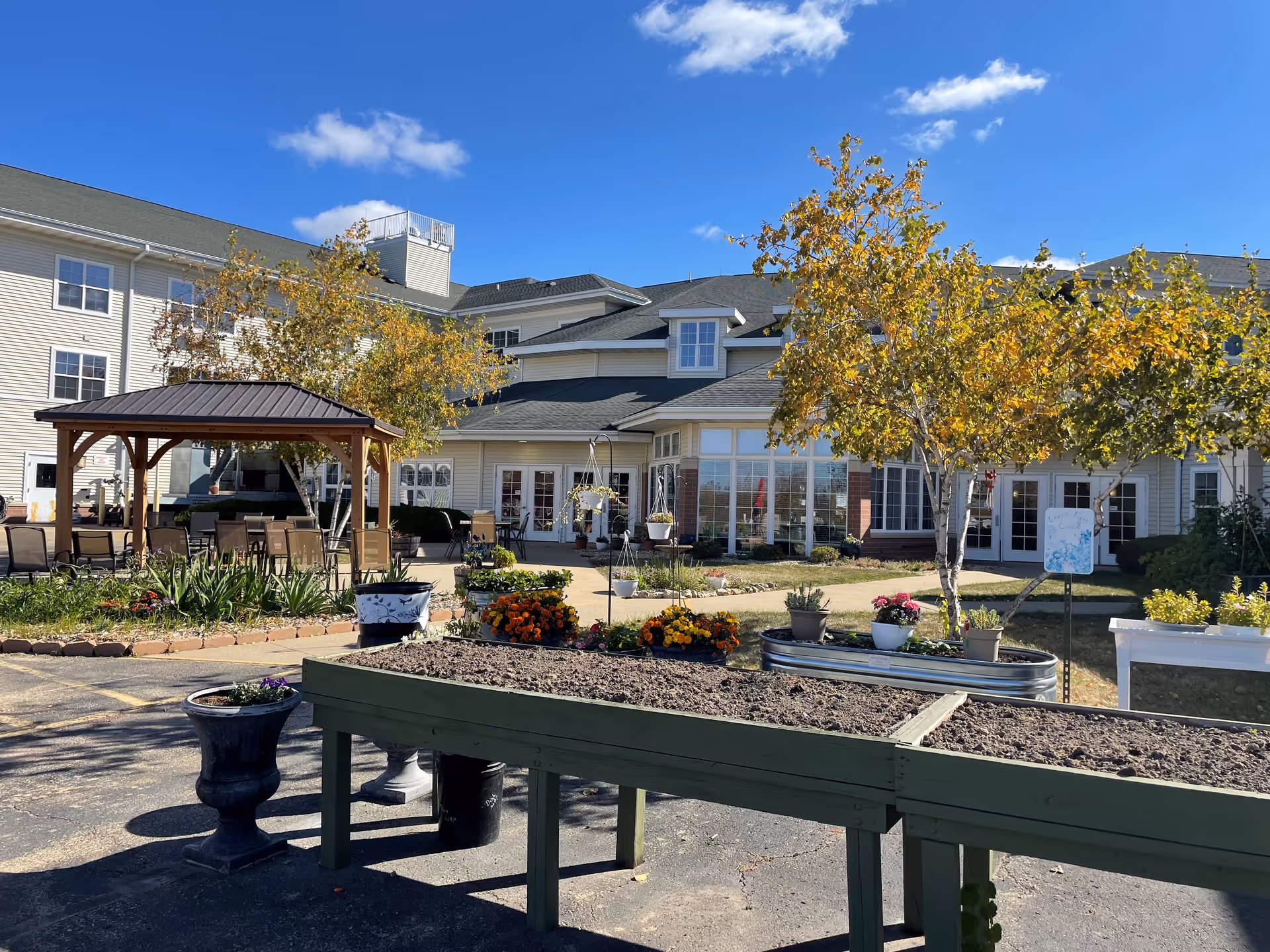 Courtyard with a gazebo, raised planter beds, flowers and trees in front of a multi-story senior living building under a blue sky.