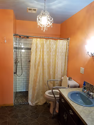 Bathroom with peach-colored walls, a blue sink on a countertop with dark cabinets, a toilet with a safety grab bar, a shower area with white subway tiles and a patterned shower curtain, and a chandelier-style light fixture on the ceiling.