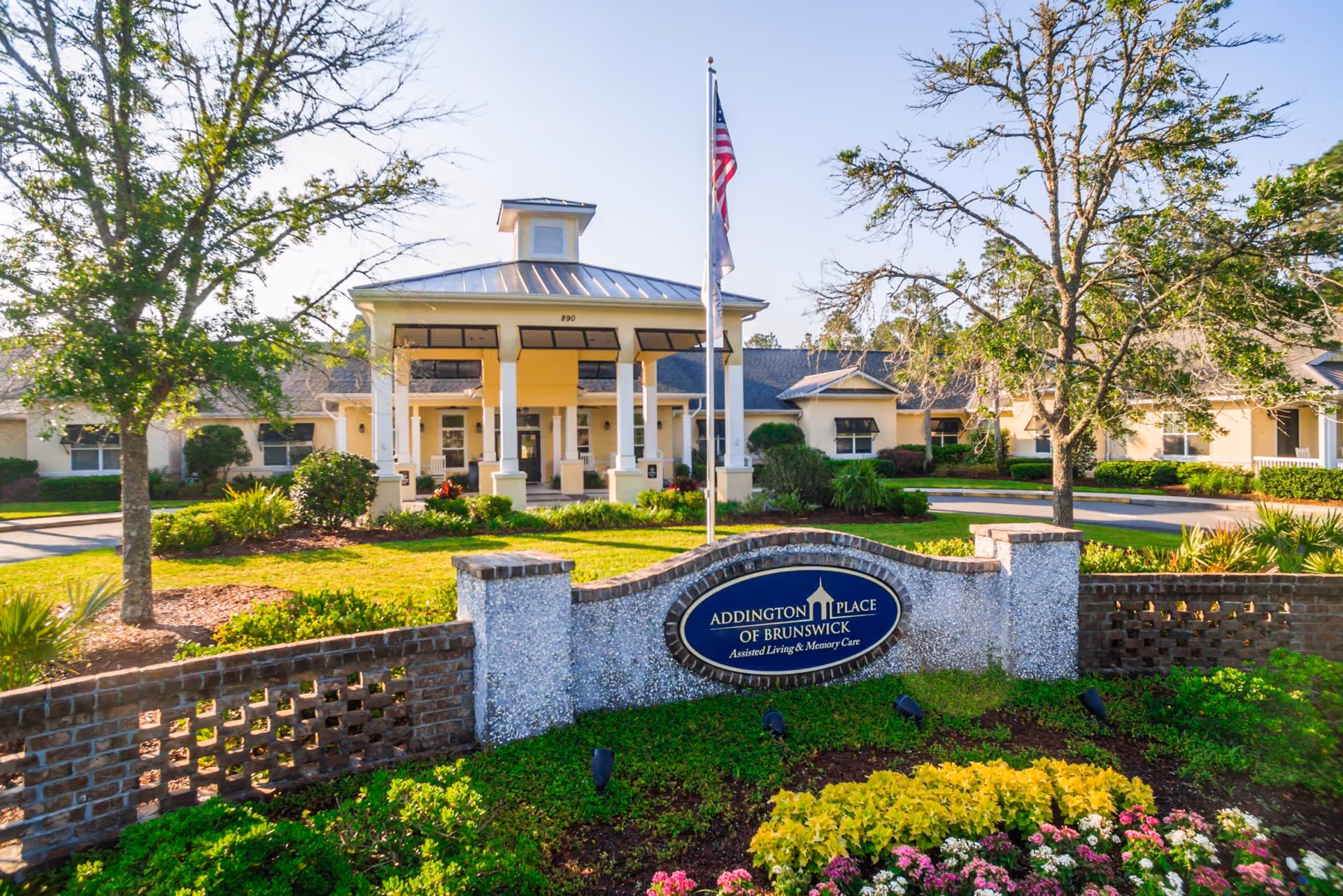 Front exterior view of Addington Place of Brunswick, an assisted living and memory care facility, featuring a well-maintained garden with colorful flowers, a brick and stone sign, a flagpole with an American flag, and a covered entrance with white columns.