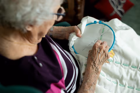 An elderly woman with gray hair and glasses is sitting and doing embroidery on a white fabric held in a blue embroidery hoop. She is wearing a purple and pink striped top.