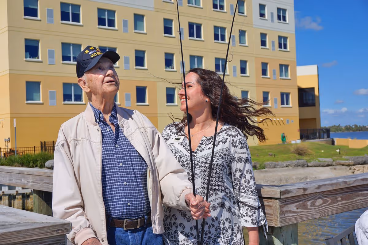 An elderly man wearing a WW II veteran cap and a light jacket stands on a wooden dock holding a fishing rod. A woman with long dark hair, wearing a patterned white and black blouse, stands beside him smiling. Behind them is a multi-story yellow and beige building with many windows, and a body of water is visible to the right under a blue sky with some clouds.