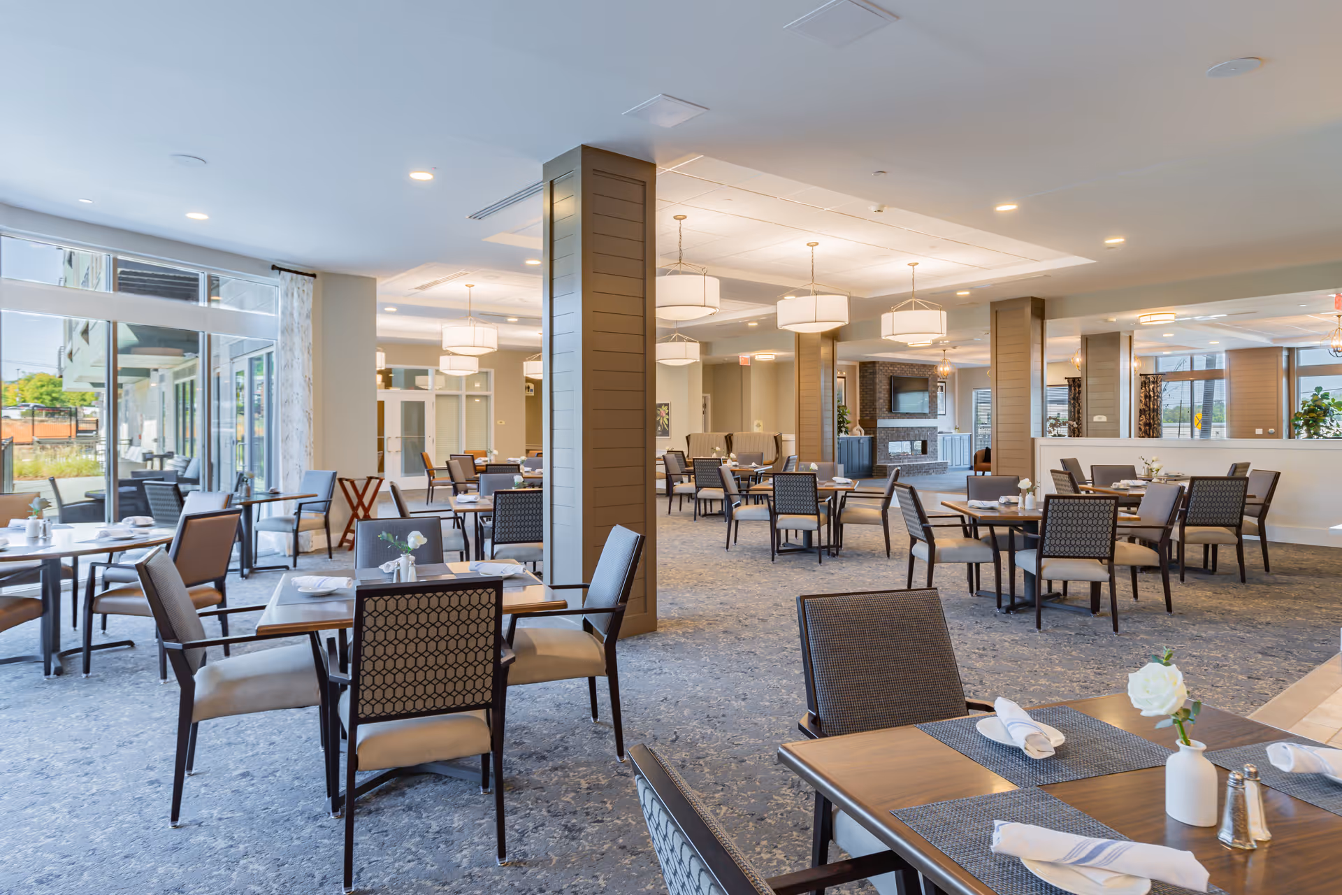 A spacious and well-lit dining room with multiple tables and chairs arranged neatly. Each table is set with placemats, napkins, and small flower vases. Large windows on the left side allow natural light to fill the room, and there are decorative light fixtures hanging from the ceiling. The room has a modern and comfortable ambiance.