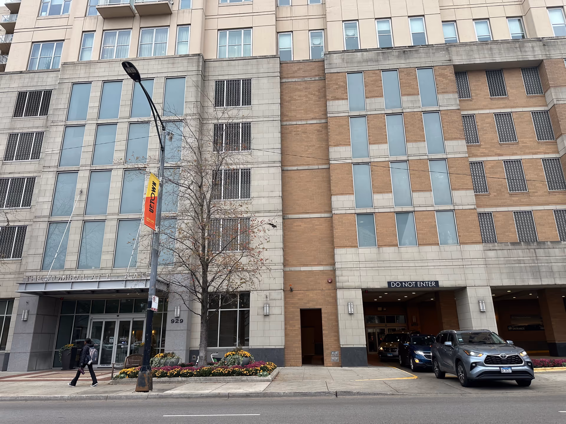 Street view of the exterior of a multi-story building named The Admiral at the Lake. The building has large windows and a mix of beige and brown brick facade. There is a sidewalk with a person walking, a tree without leaves, flower beds, and a streetlamp with a banner. Several cars are visible entering or exiting a parking garage with a 'DO NOT ENTER' sign above one entrance.