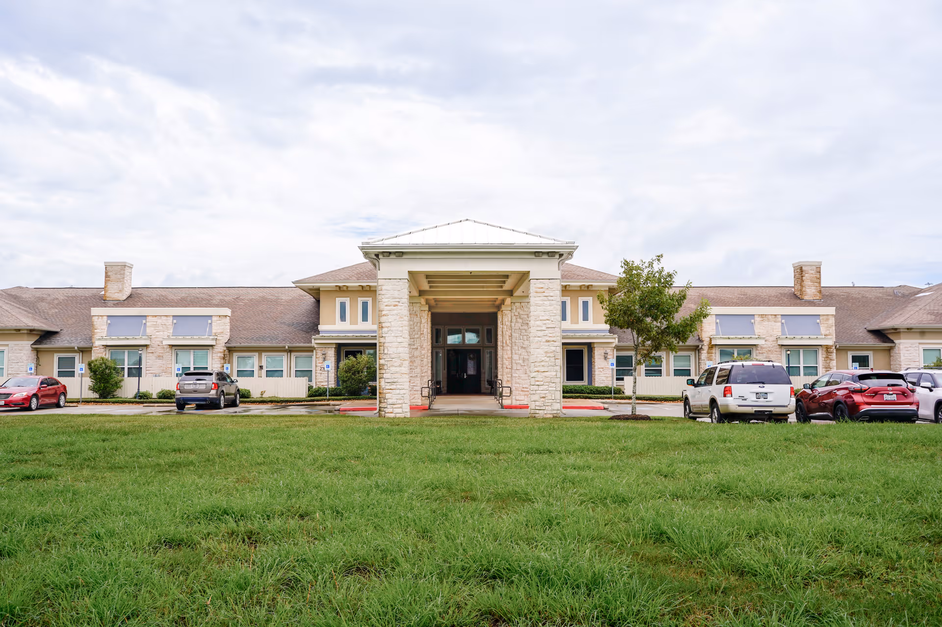 Front exterior view of Orchard Park at Victory Lakes Assisted & Senior Living building with a covered entrance, stone pillars, several windows, a grassy lawn in the foreground, and parked cars on either side.