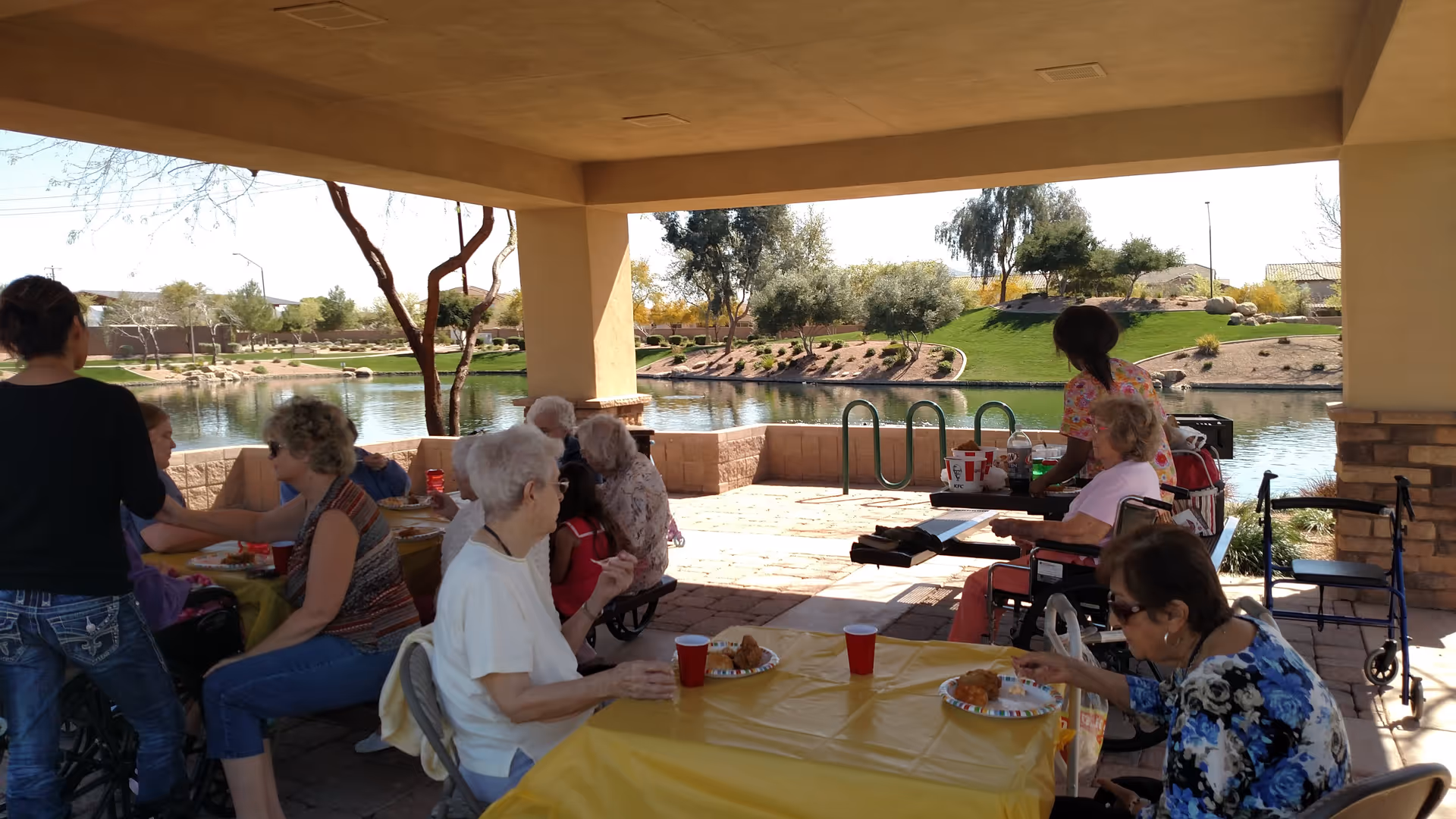 A group of elderly people and caregivers gathered under a covered outdoor patio near a pond, sitting at tables with yellow tablecloths and eating food from plates. The background shows a landscaped area with grass, trees, and shrubs.
