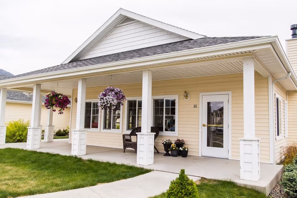 Exterior view of a single-story beige building with a covered porch supported by white columns. The porch has hanging flower baskets and potted plants near the entrance door marked with the number 26. There is a wicker chair on the porch and a well-maintained lawn in front.