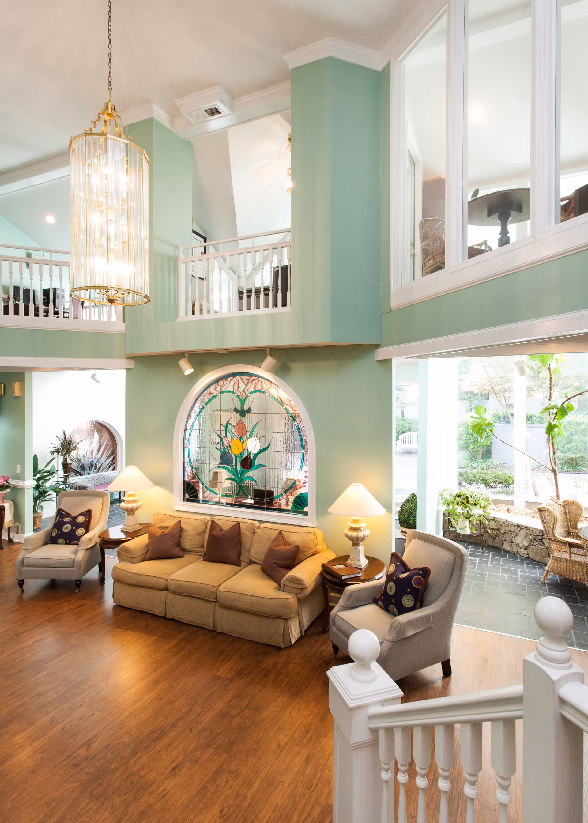 A bright and spacious living room area with light green walls and wooden flooring. The room features a beige sofa with brown cushions, two armchairs with patterned pillows, two side tables with lamps, a large decorative stained glass window, and a large hanging chandelier. There is a staircase with white railings in the foreground and large windows looking out to a garden patio area with wicker chairs and plants.