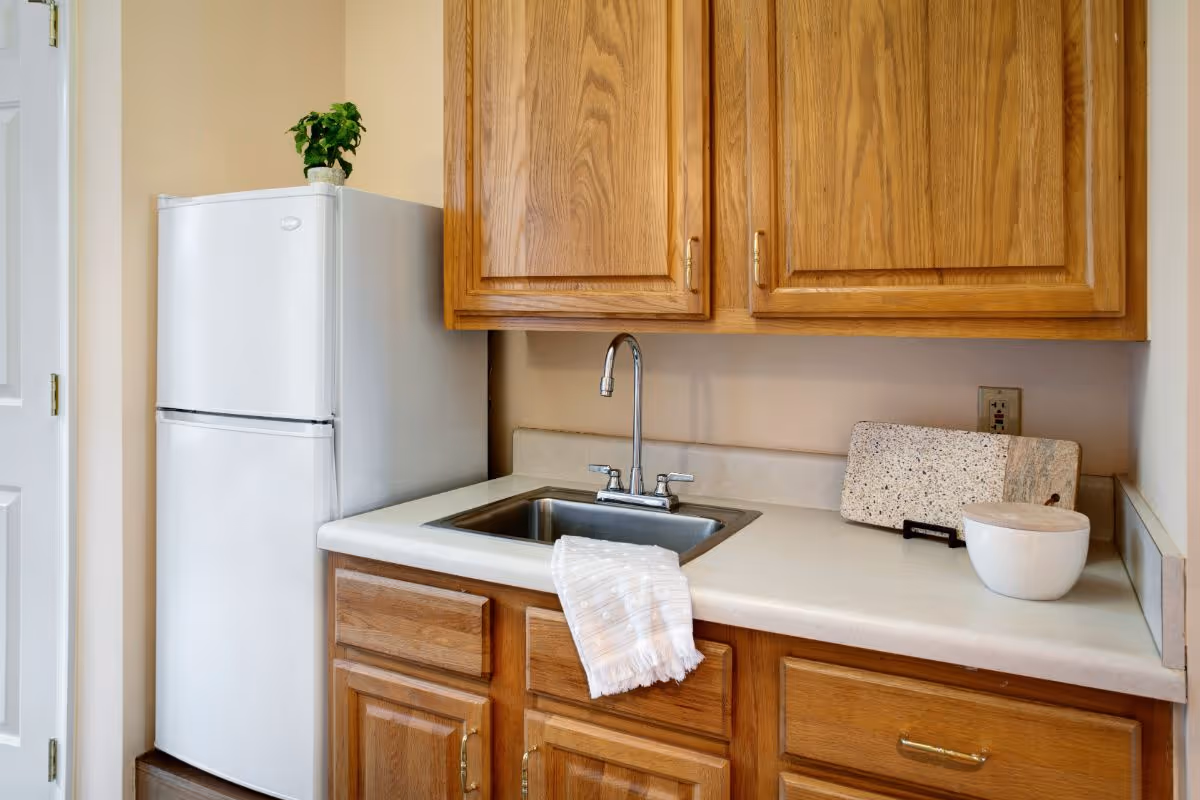 A kitchen area with wooden cabinets, a white countertop, a stainless steel sink with a faucet, a white refrigerator, a small potted plant on top of the refrigerator, a white bowl, and a cutting board on the countertop.