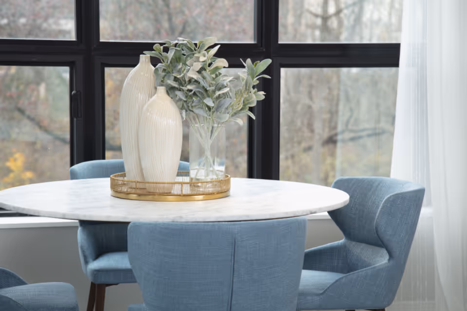 A round white marble table with a gold tray holding two tall cream-colored vases and a clear vase with green leafy branches. The table is surrounded by four blue upholstered chairs. Large windows with black frames and sheer white curtains provide a view of trees outside.