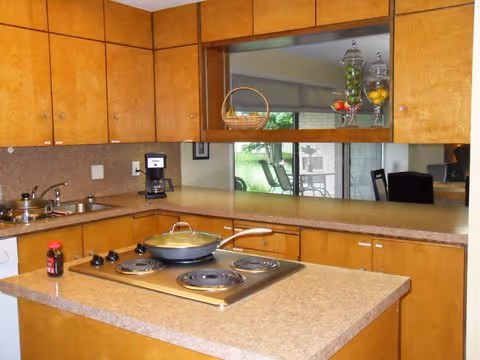 Small kitchen with wood cabinets, a countertop island with an electric cooktop, sink and coffee maker, and a pass-through window to a dining area.