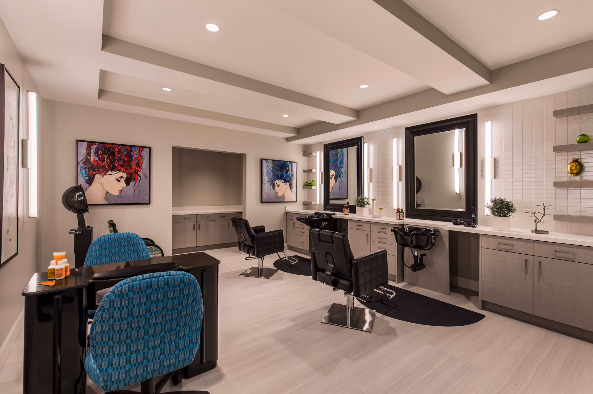 A modern salon area inside a senior living facility with two black salon chairs in front of large mirrors and sinks for hair washing. There are two blue patterned chairs at a black manicure table with nail polish bottles on it. The walls are decorated with colorful paintings of women's faces with vibrant hair. The room has light wood flooring, recessed lighting, and gray cabinetry with plants and decorative items on the counters and shelves.