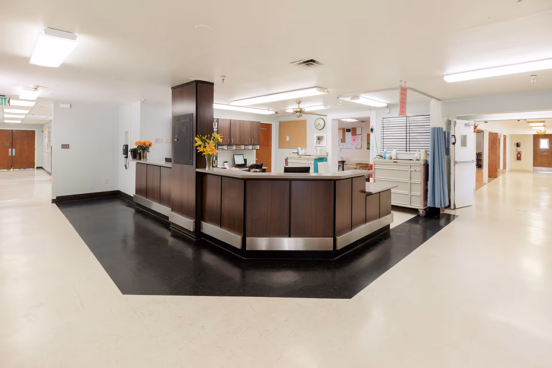 A clean and well-lit reception desk area in a senior care facility with dark wood paneling and a black floor section around it. The surrounding hallway has light-colored floors and walls, with doors and bulletin boards visible in the background. There are flowers placed on the reception desk, and fluorescent ceiling lights illuminate the space.