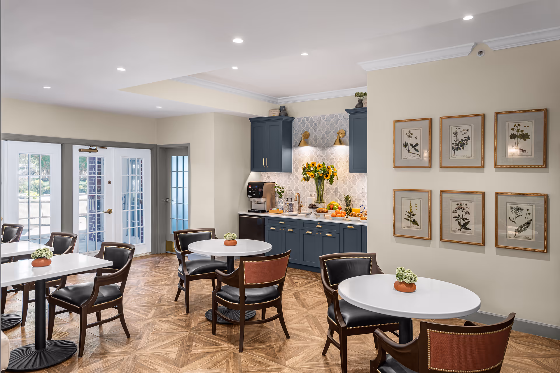A bright dining area with several round and rectangular tables surrounded by dark wooden chairs with leather seats. The room features a small kitchenette with blue cabinets, a coffee machine, a vase of sunflowers, and a fruit display. The walls are decorated with framed botanical prints, and large windows and glass doors allow natural light to enter.