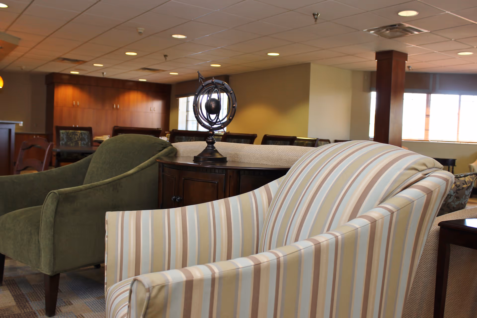Interior view of a senior living facility lounge area with a striped armchair in the foreground, a green armchair beside it, a wooden cabinet with a decorative globe sculpture on top, and dining tables and chairs in the background under a ceiling with recessed lighting.