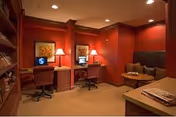 A cozy interior room with warm red walls featuring two computer desks with chairs, each desk having a lamp and framed artwork above. There is a small seating area with a cushioned bench and a round wooden table. The room is softly lit with recessed ceiling lights.