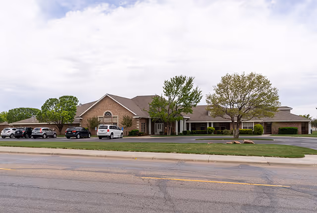 Exterior view of a single-story brick building with a pitched roof, several trees, and a parking lot with multiple cars parked in front. The sky is partly cloudy.