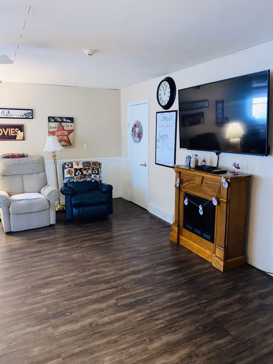 A cozy living room area with two armchairs, one beige and one dark blue with a dog-themed blanket draped over it. There is a floor lamp between the chairs, wall decorations including framed signs, a clock, and a whiteboard. A large flat-screen TV is mounted above a wooden electric fireplace on a wood floor.