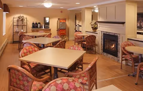 A cozy common area with several square tables surrounded by cushioned chairs featuring a red and beige patterned upholstery. There is a fireplace with a fire burning on the right side of the room, and a beverage station with coffee dispensers and a popcorn machine against the back wall. The room has wooden flooring and soft lighting from ceiling fixtures.