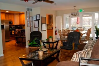 Interior view of a senior living facility showing a living room area with cushioned chairs and a coffee table with a plant, adjacent to a dining room with a wooden dining table and chairs. The kitchen is visible in the background with wooden cabinets and stainless steel appliances. Large windows and a glass door provide natural light and a view of an outdoor patio.