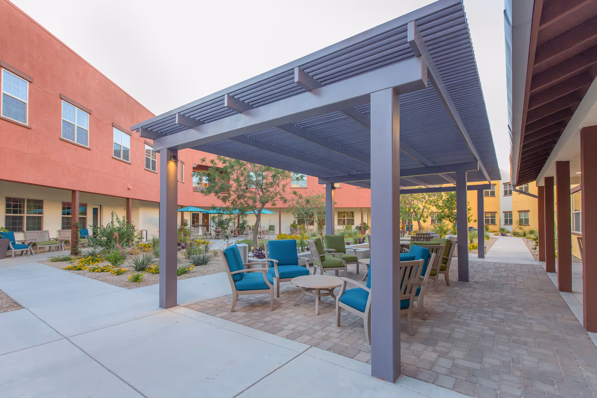Outdoor courtyard area at The Ranch Estates of Tucson featuring a shaded pergola with several cushioned chairs and small tables underneath. Surrounding the pergola are walkways, landscaped garden beds with flowers and shrubs, and multi-story buildings with windows and balconies.
