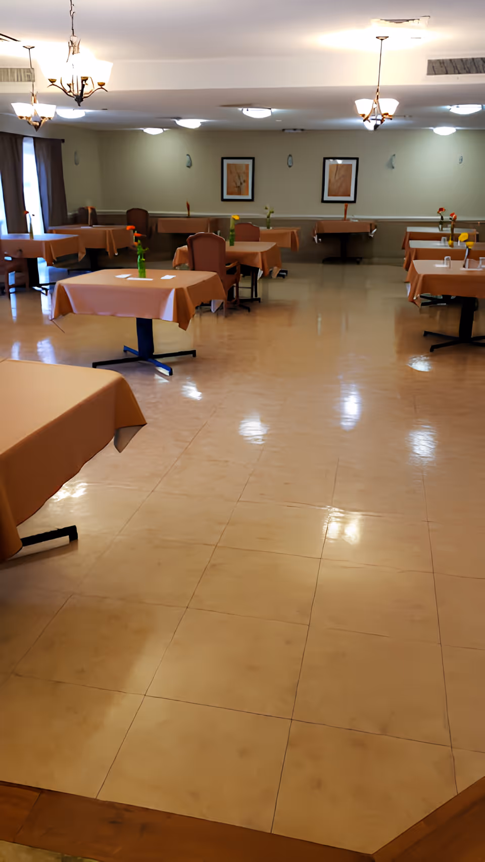Spacious dining room with multiple tables covered in beige tablecloths, chairs, small vases with flowers, and overhead chandeliers.