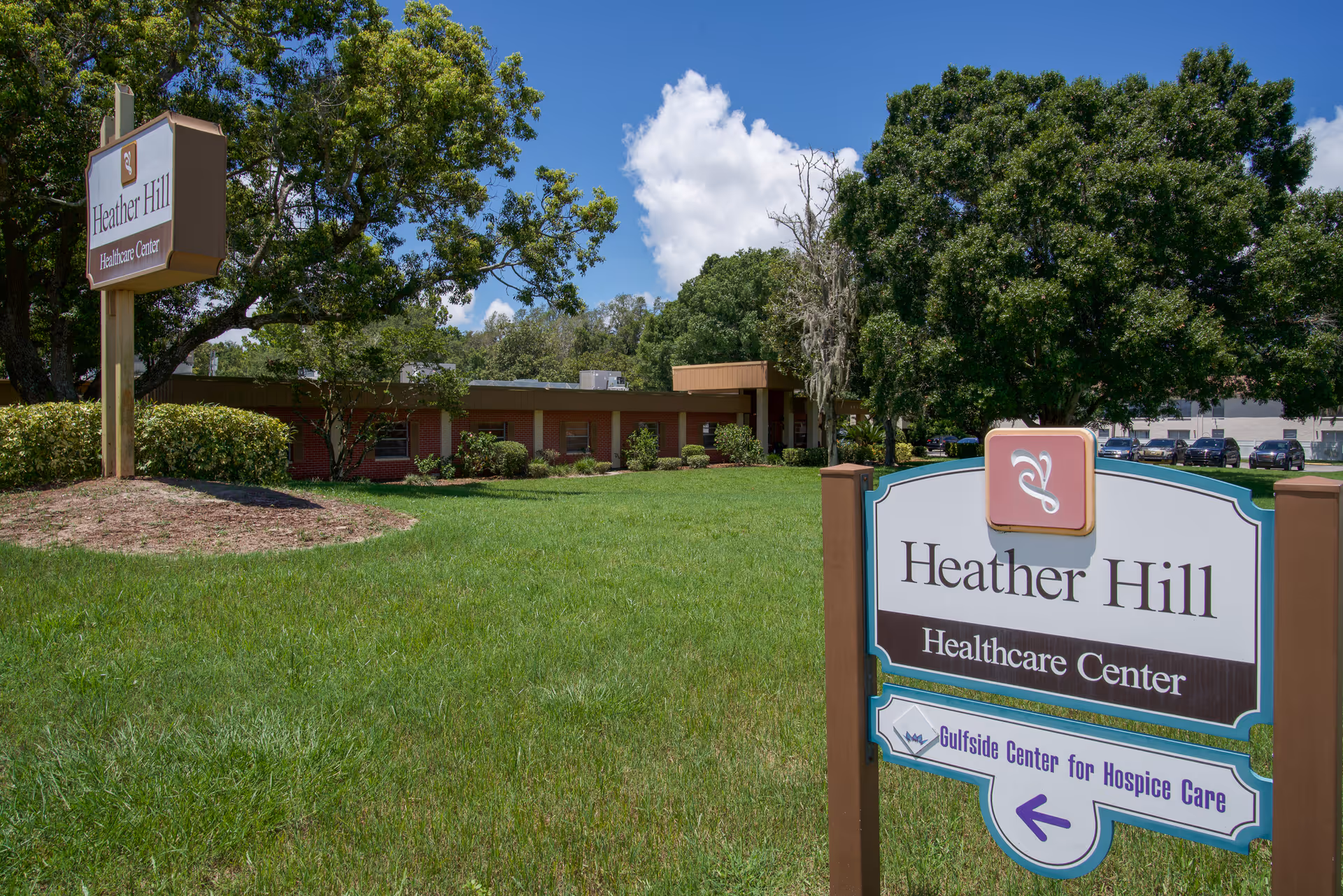 Exterior view of Heather Hill Healthcare Center with a large green lawn, trees, and two signs displaying the facility's name. The sky is blue with some clouds, and there are parked cars visible in the background.