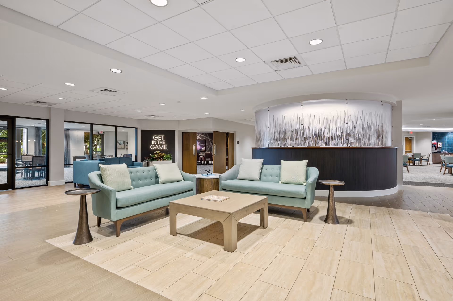 A modern senior living facility lounge area with two light blue sofas facing each other, each with two light-colored pillows. Between the sofas is a light wooden coffee table, and on each side of the sofas is a small round side table. In the background, there is a curved reception desk with a decorative panel above it. The space has light-colored tile flooring and a drop ceiling with recessed lighting. Large windows and glass doors are visible on the left side, allowing natural light to enter. A sign on the wall reads 'GET IN THE GAME'.