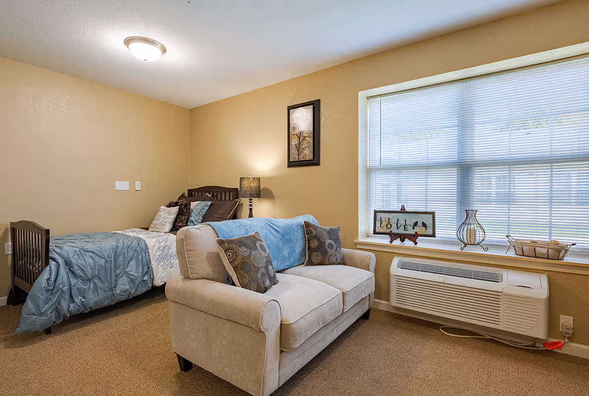 A cozy bedroom in Chandler Place featuring a single bed with blue and white bedding, a beige loveseat with decorative pillows, a lamp on a small table, a framed picture on the wall, and a large window with blinds above a heating and air conditioning unit. The room has beige walls and carpeted flooring.
