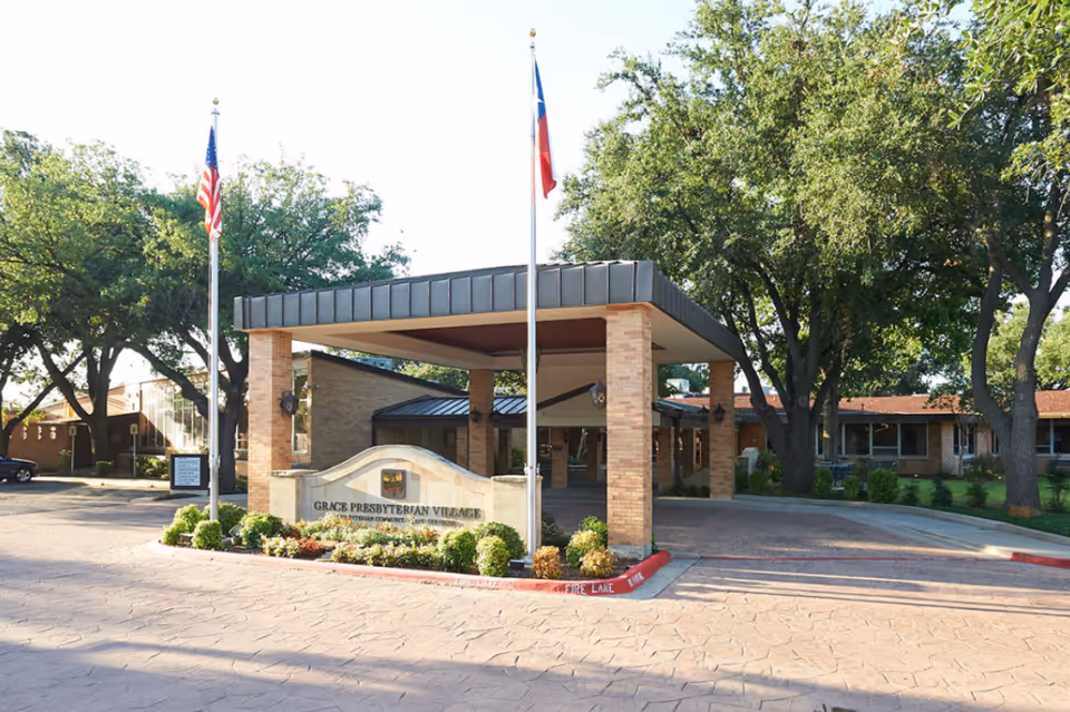 Entrance of Grace Presbyterian Village senior living facility with a covered drop-off area supported by brick pillars, two flagpoles with American and Texas flags, surrounded by trees and landscaping.