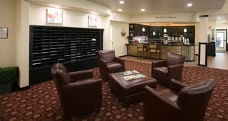 A cozy seating area in a senior living facility with four brown leather armchairs arranged around a square ottoman with magazines on top. Behind the seating area is a wall with multiple mail slots and framed artwork above. To the right, there is a reception desk with a stone facade and pendant lights hanging above. The carpet is patterned in shades of red and brown, and the walls are painted beige.