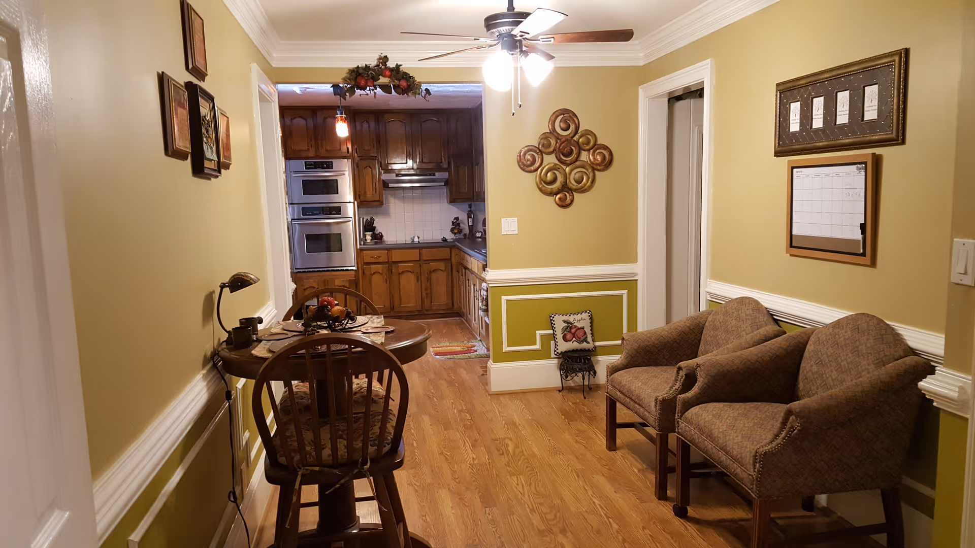 Interior view of a cozy living space with two upholstered armchairs on the right, a small round dining table with two wooden chairs on the left, and a kitchen area with wooden cabinets and double ovens in the background. The walls are painted yellow with white trim, decorated with framed pictures and wall art. A ceiling fan with lights is mounted above.