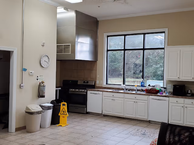 A clean kitchen area with white cabinets, a large window showing outdoor greenery, a stainless steel stove with a vent hood, a dishwasher, a sink with dishes, and various kitchen appliances on the countertop. There are three trash bins and a yellow caution wet floor sign near the entrance, along with a wall clock and a fire extinguisher mounted on the wall.