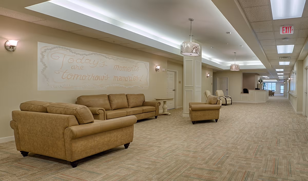 Spacious, well-lit senior living lounge with beige sofas and armchairs, decorative wall lettering, pendant lights, and a long carpeted corridor leading to a reception area.
