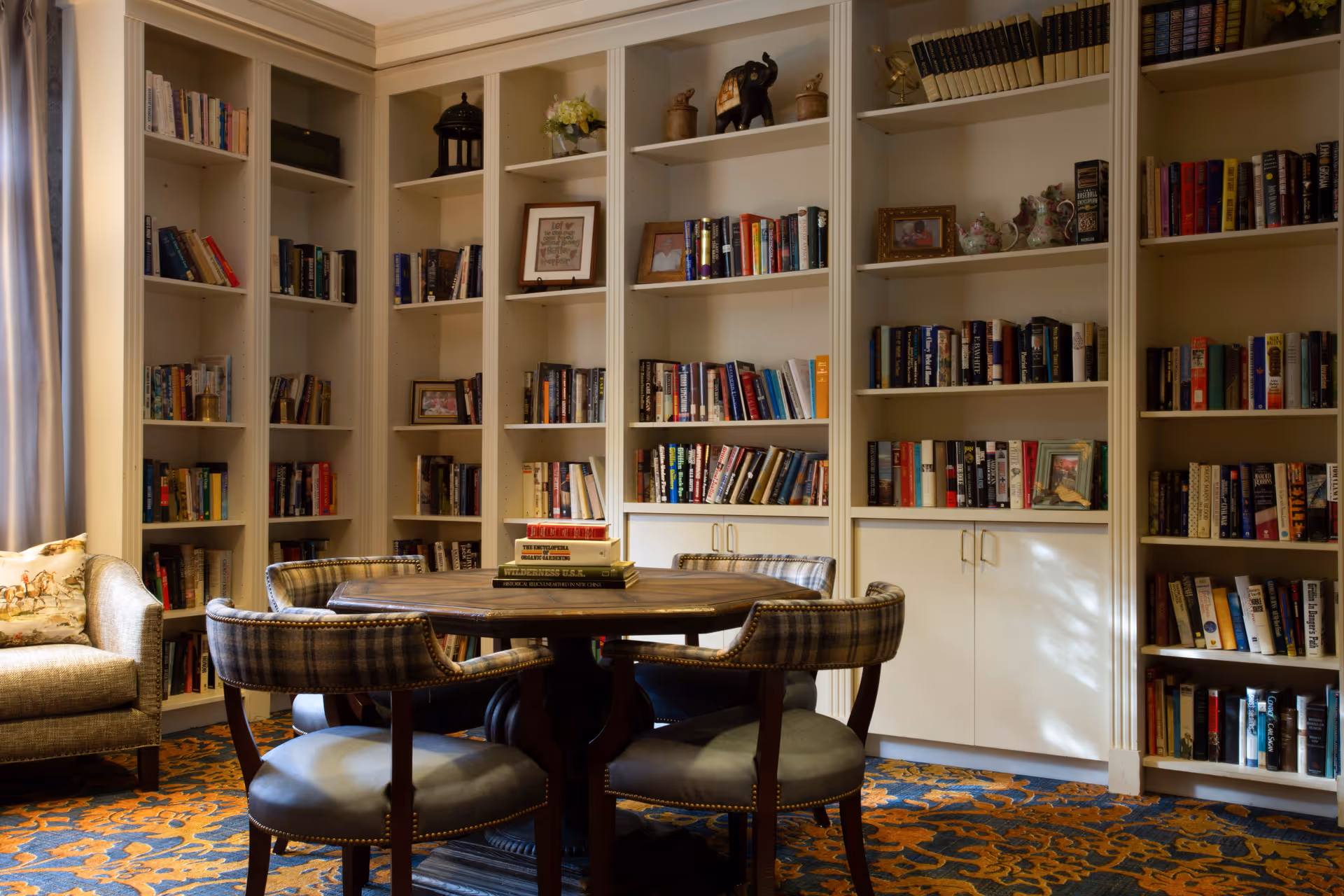 A cozy library room with built-in white bookshelves filled with books and decorative items. In the center, there is a round wooden table surrounded by four upholstered chairs with plaid backs and cushioned seats. To the left, there is a cushioned armchair next to a window with curtains. The floor is covered with a patterned carpet in blue and orange tones.