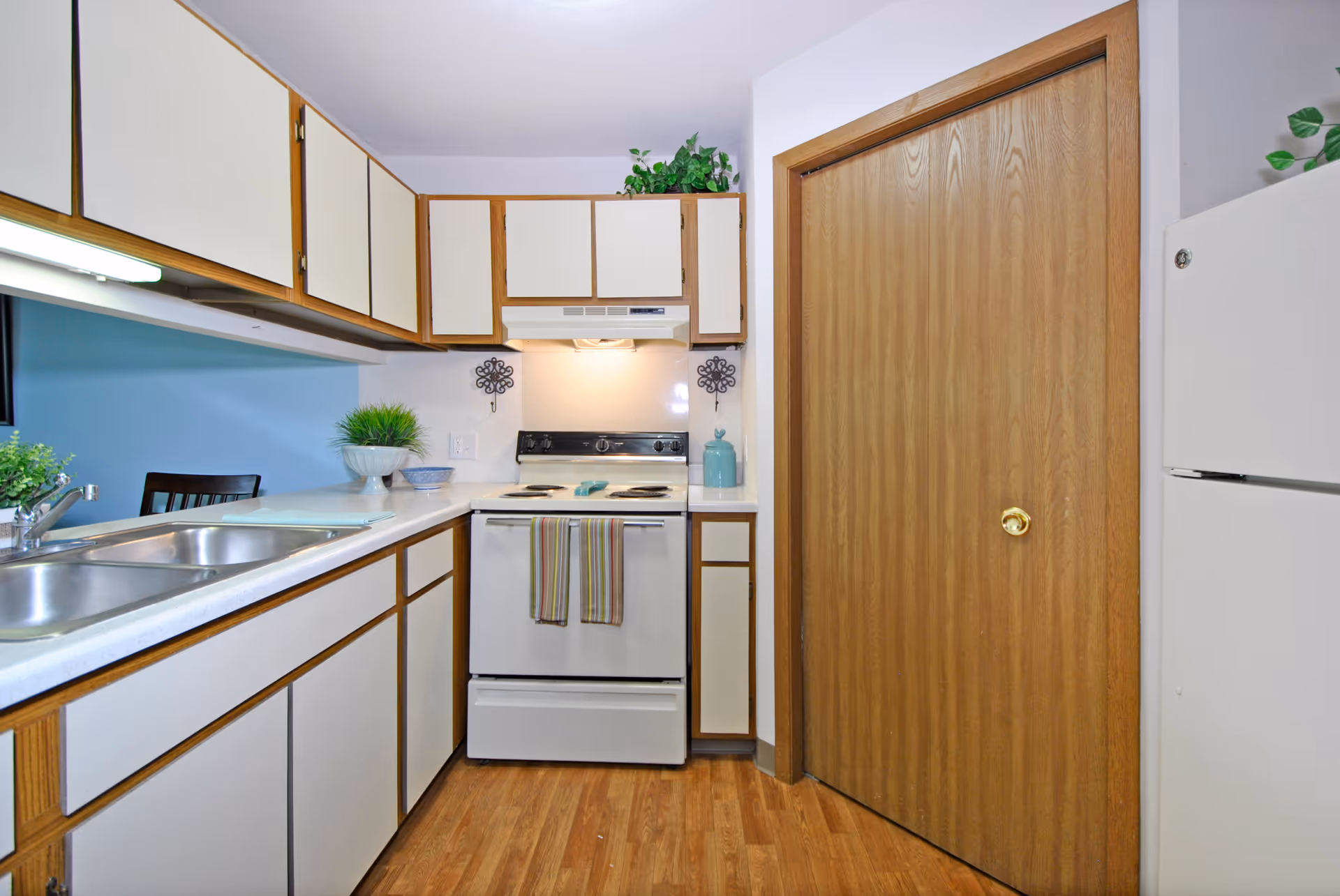 A compact kitchen with white cabinets and wooden trim, a white electric stove with two striped towels hanging on the handle, a double stainless steel sink, a white refrigerator, and a wooden pantry door. There are small decorative plants on the countertop and above the cabinets.