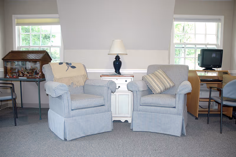 A cozy sitting area with two blue and white checkered armchairs, one with a beige throw blanket and the other with a striped pillow, positioned on a carpeted floor. Between the chairs is a small white cabinet with a dark blue lamp on top. Behind the chairs are two windows letting in natural light. To the left, there is a table with a dollhouse, and to the right, a desk with a computer and chair.