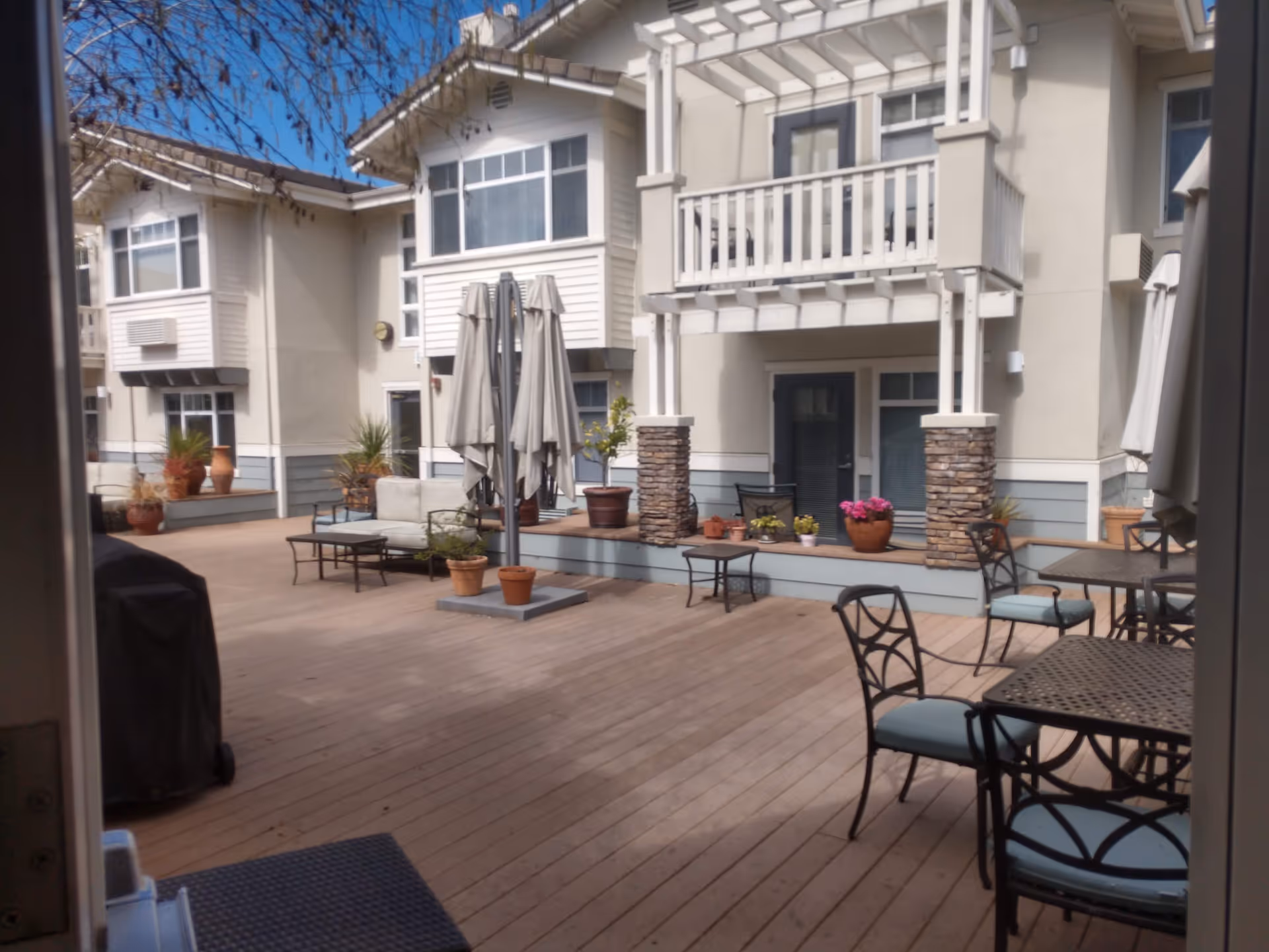 Outdoor patio area at Fremont Hills featuring wooden decking, several metal tables and chairs with cushions, potted plants, closed umbrellas, and a two-story building with balconies and windows in the background under a clear blue sky.