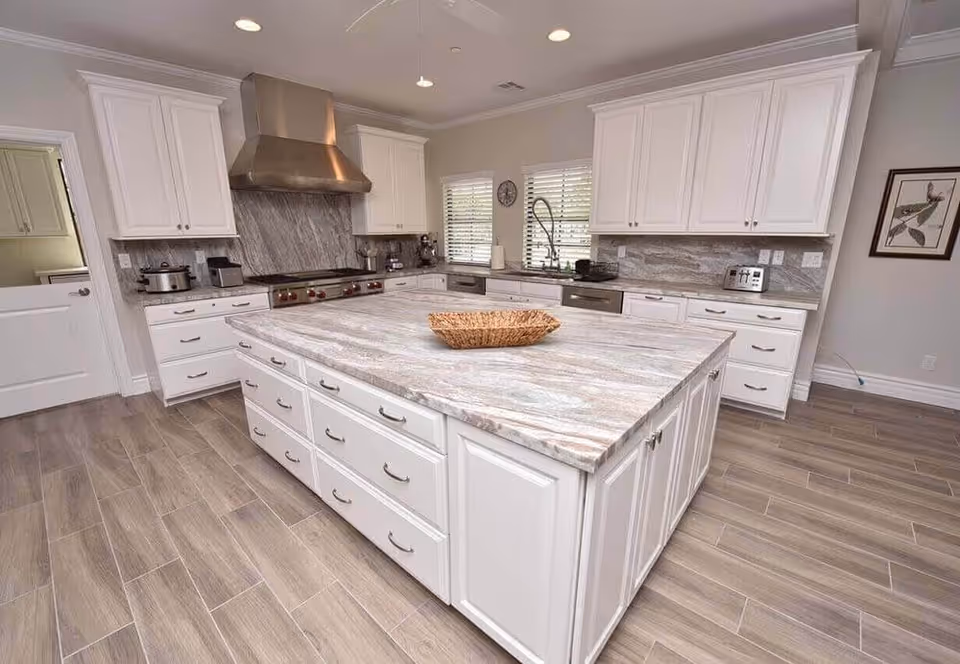 A spacious modern kitchen with a large marble island countertop in the center featuring multiple drawers and cabinets. The kitchen has white cabinetry, stainless steel appliances including a large range hood, stove, and dishwasher. There are two windows with blinds above the sink, a ceiling fan, and a framed picture on the wall.